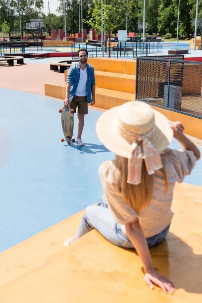 Усміхнений чоловік тримає довгу дошку біля розмитої дівчини в солом'яному капелюсі в скейт-парку — стокове фото