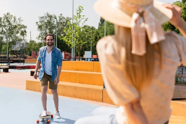 Hombre alegre montando longboard cerca de novia borrosa en skate park - foto de stock