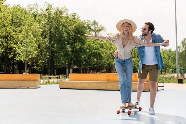 Mulher positiva montando longboard perto namorado em óculos de sol no parque de skate — Fotografia de Stock