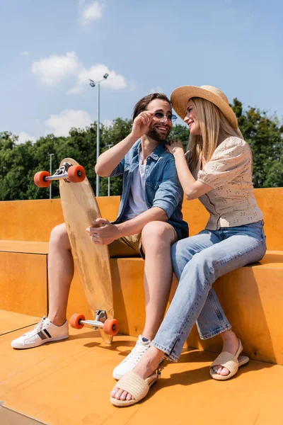 Smiling couple in sunglasses and sun hat holding longboard in skate park — Stock Photo