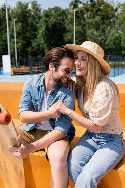 Pareja positiva cogida de la mano cerca de longboard en skate park - foto de stock