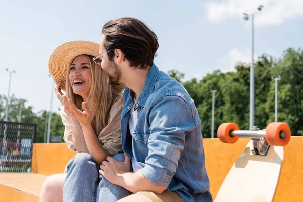 Fröhliche Frau mit Sonnenhut unterhält sich mit Freund nahe Longboard im Skatepark — Stockfoto