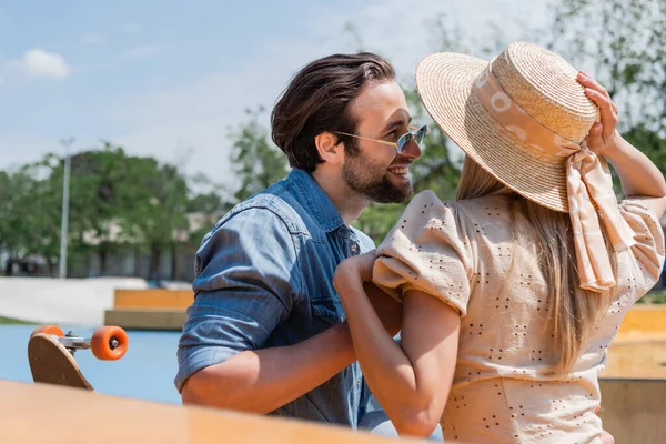 Cheerful man in sunglasses looking at girlfriend in straw hat near longboard in skate park — Stock Photo