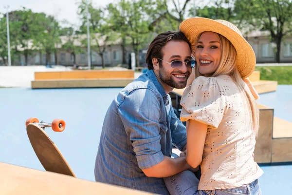 Pareja positiva mirando la cámara cerca de longboard en skate park - foto de stock