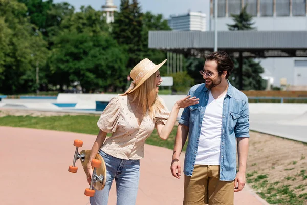 Fröhliche Frau mit Sonnenhut hält Longboard in der Hand und spricht mit Freund im Skatepark — Stockfoto
