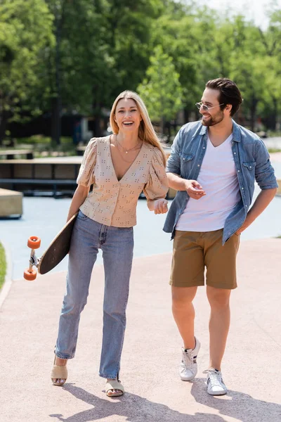 Pareja feliz con longboard caminando en skate park durante el día - foto de stock