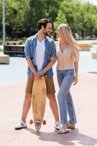 Side view of man in sunglasses holding longboard near smiling girlfriend in skate park — Stock Photo