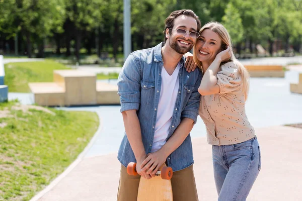 Pareja positiva con longboard mirando a la cámara en skate park - foto de stock