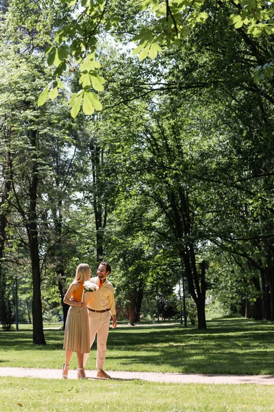 Smiling woman holding bouquet while walking near boyfriend in summer park — Stock Photo