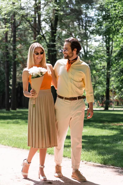 Elegante donna che tiene bouquet vicino al fidanzato sorridente con occhiali da sole nel parco — Foto stock