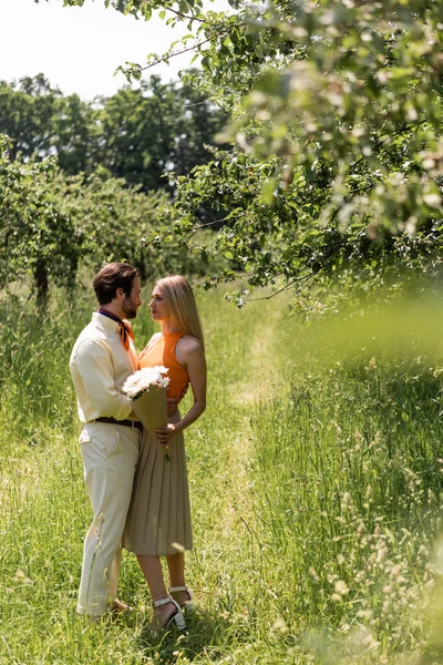 Side view of stylish woman holding bouquet and looking at boyfriend in park — Stock Photo