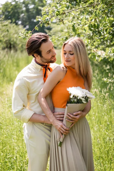 Young man hugging smiling girlfriend with bouquet in summer park — Stock Photo