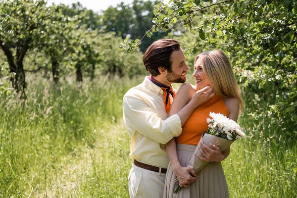 Cheerful man touching stylish girlfriend with bouquet in park — Stock Photo