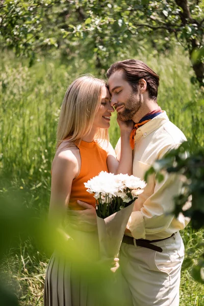 Vista laterale della donna sorridente che tiene il bouquet e tocca il fidanzato elegante nel parco — Foto stock