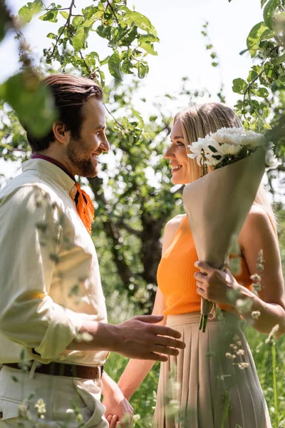 Seitenansicht des lächelnden stilvollen Paares mit Blumenstrauß Händchen haltend im Sommerpark — Stockfoto