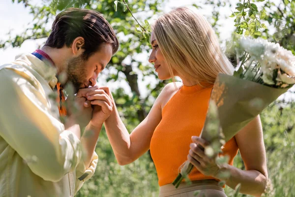 Vista laterale di uomo elegante baciare la mano della fidanzata tenendo bouquet nel parco — Foto stock