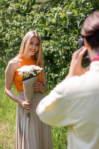 Lächelnde Frau hält Blumenstrauß neben verschwommenem Freund mit Oldtimer-Kamera im Park — Stockfoto