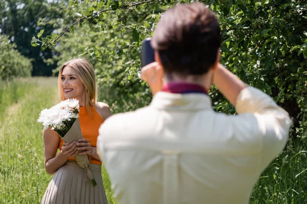 Mujer positiva con el ramo posando cerca borrosa novio con cámara retro en el parque - foto de stock