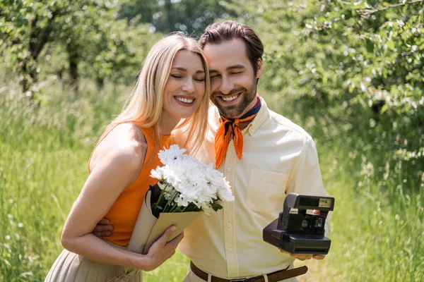 Smiling and trendy couple holding retro camera and bouquet while hugging in summer park — Stock Photo