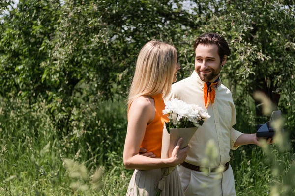 Elegante uomo che tiene fotocamera vintage e abbracciando la ragazza con bouquet nel parco estivo — Foto stock