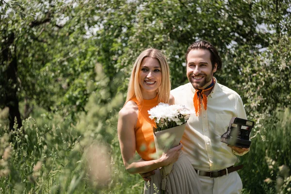 Stylish couple with bouquet and retro camera looking away in summer park — Stock Photo