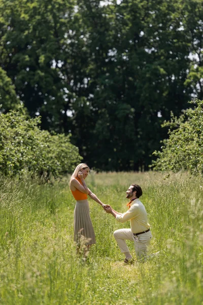 Side view of stylish man kneeling and holding hand of girlfriend on meadow in park — Stock Photo