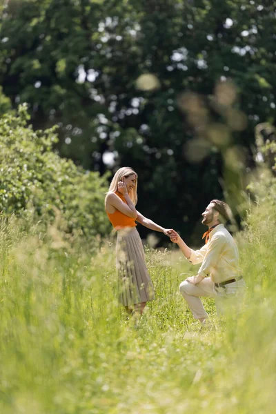 Vista lateral do homem na moda segurando a mão da namorada e ajoelhado no gramado no parque — Fotografia de Stock