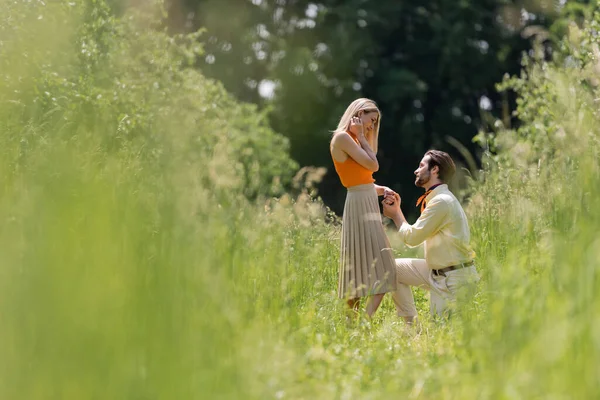 Seitenansicht eines stilvollen Mannes, der die Hand seiner Freundin hält, während er im Sommerpark kniet — Stockfoto