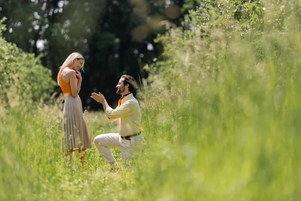 Side view of smiling man pointing at stylish girlfriend while kneeling on lawn in park — Stock Photo