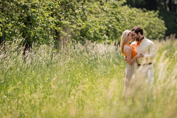 Вид сбоку улыбающейся пары, держащейся за руки и обнимающейся на лужайке в летнем парке — стоковое фото