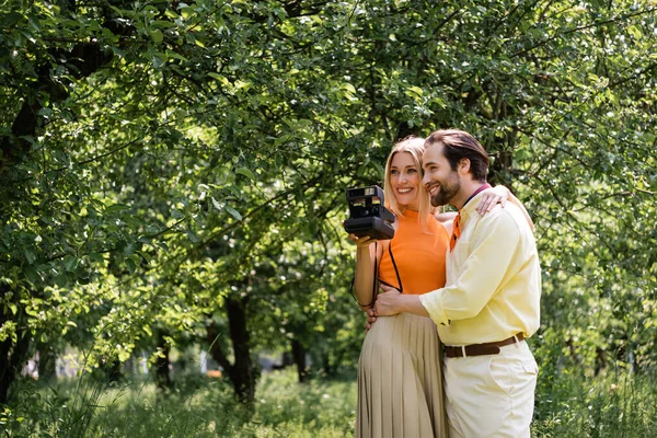 Sorrindo homem abraçando namorada elegante com câmera vintage no parque de verão — Fotografia de Stock