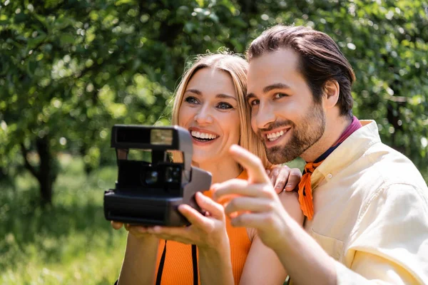 Elegante hombre apuntando con el dedo cerca de la novia con la cámara retro borrosa en el parque - foto de stock