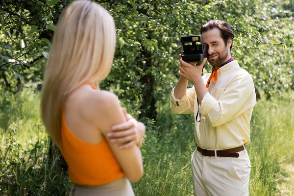 Homem na moda tirando foto na câmera vintage perto de namorada borrada no parque de verão — Fotografia de Stock