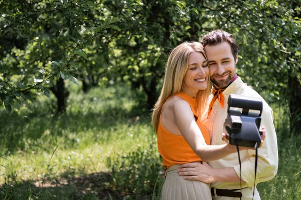 Sonriente y de moda pareja tomando fotos en la cámara retro en el parque de verano - foto de stock