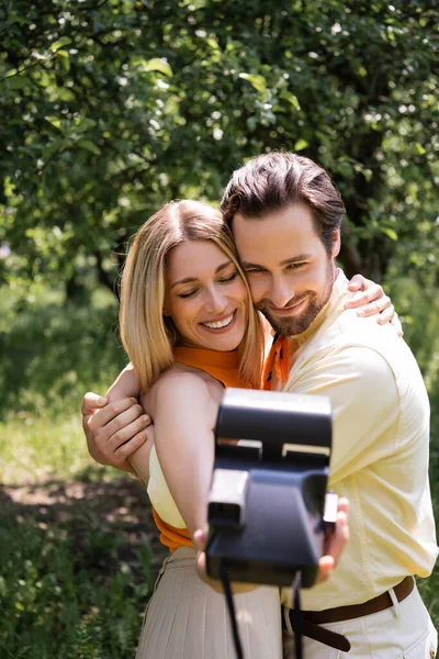 Stylish couple embracing while taking photo on retro camera in park — Stock Photo