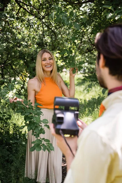 Femme élégante debout près de l'arbre près du petit ami flou avec caméra rétro dans le parc — Photo de stock