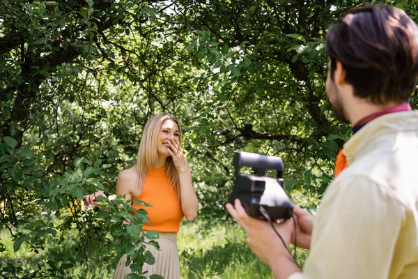 Donna allegra in piedi vicino all'albero e fidanzato sfocato con fotocamera vintage nel parco — Foto stock