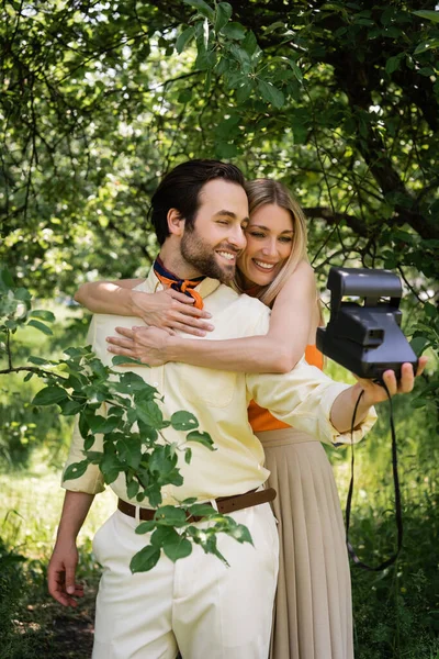 Smiling woman embracing stylish boyfriend taking photo on retro camera in summer park — Stock Photo