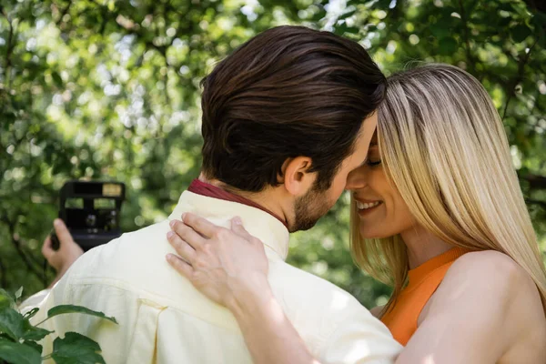 Smiling woman hugging boyfriend with blurred retro camera in summer park — Stock Photo