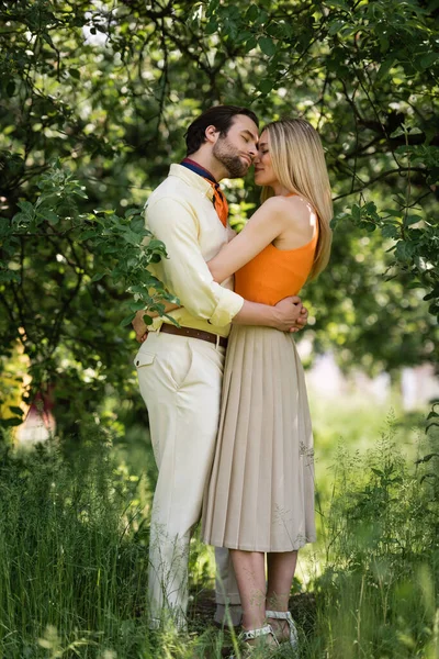 Vista lateral de pareja elegante abrazando cerca de los árboles en el parque de verano - foto de stock