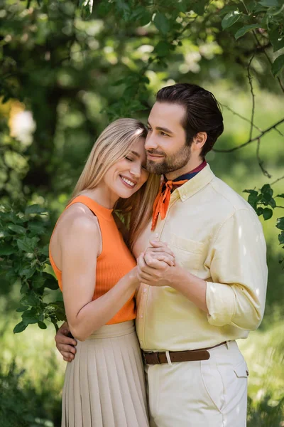 Stylish man hugging and holding hand of smiling girlfriend in park — Stock Photo