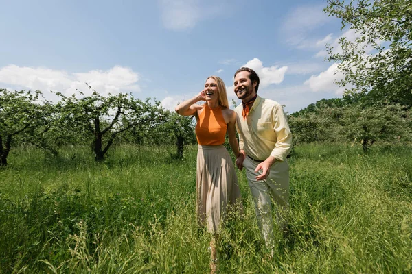 Femme à la mode tenant la main du petit ami souriant tout en marchant dans le parc d'été — Photo de stock