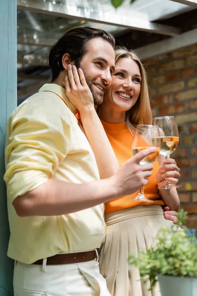 Mujer con estilo tocando la cara de novio sonriente con vino en la cafetería al aire libre - foto de stock