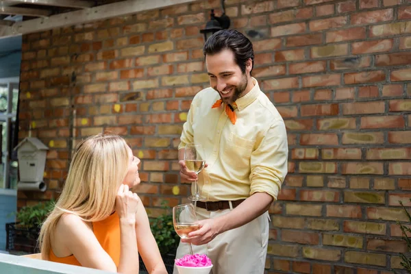Стильный мужчина с бокалами вина возле улыбающейся девушки в кафе на открытом воздухе — стоковое фото