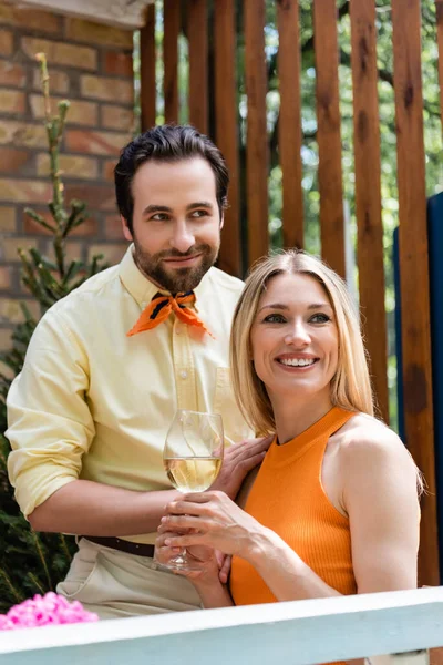 Mujer sonriente sosteniendo copa de vino cerca de novio de moda en la cafetería al aire libre - foto de stock