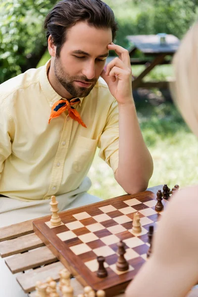 Hombre pensativo jugando ajedrez con novia borrosa en el parque - foto de stock