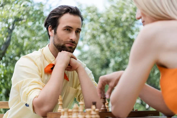 Stylish man looking at blurred girlfriend playing chess in park — Stock Photo