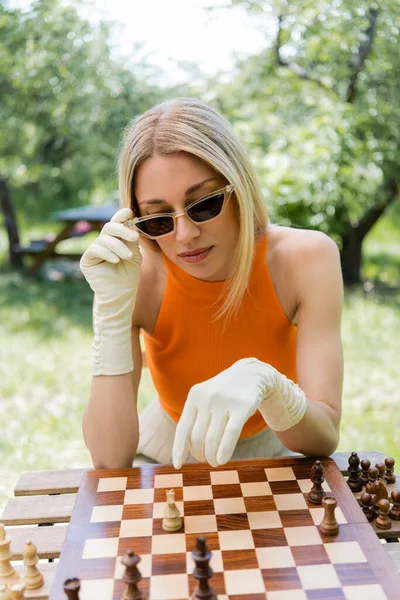 Stylish woman in sunglasses sitting near chess on board in park — Stock Photo