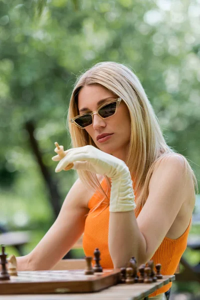 Mujer de moda sosteniendo figura de ajedrez cerca del tablero en el parque - foto de stock