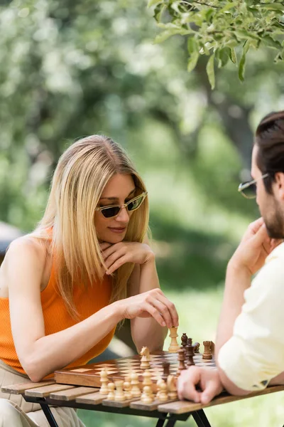 Mulher na moda jogando xadrez perto de jovem namorado borrado no parque — Fotografia de Stock